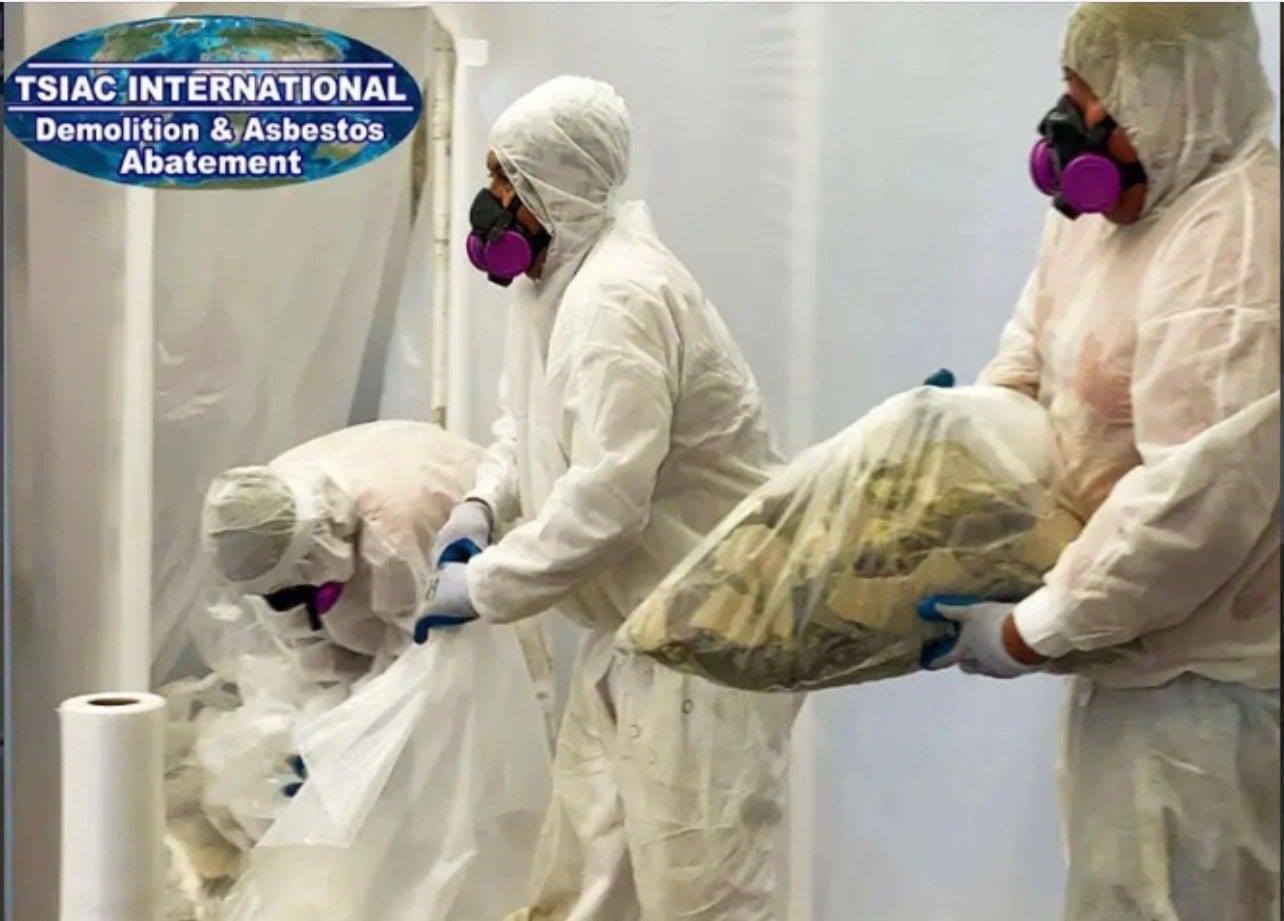 Two workers in white hazmat suits and respirators bagging asbestos debris inside a sealed work area